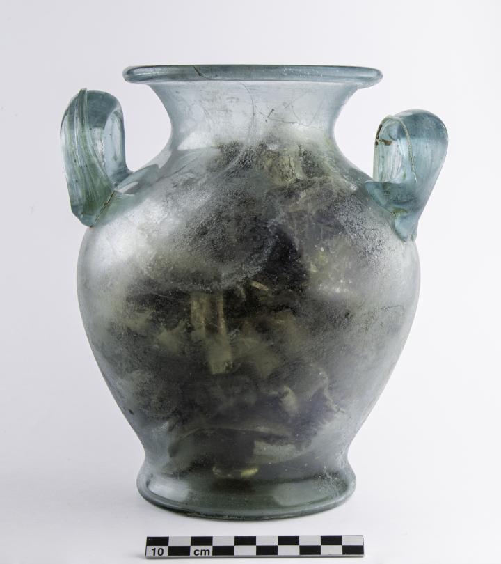 Vase ossuaire en verre contenant des fragments osseux brûlés et, dans le fond, une bague en or. Vase ossuaire en verre contenant des fragments osseux brûlés et, dans le fond, une bague en or.