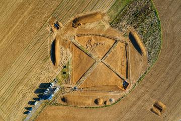 Vue zénithale de la fouille ; le tumulus apparaît, vaste aménagement de pierres et de terre ; au cœur de la fouille, à la croisée des bermes, se trouve la tombe, recoupée par la fouille de 1953.