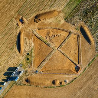 Vue zénithale de la fouille ; le tumulus apparaît, vaste aménagement de pierres et de terre ; au cœur de la fouille, à la croisée des bermes, se trouve la tombe, recoupée par la fouille de 1953.